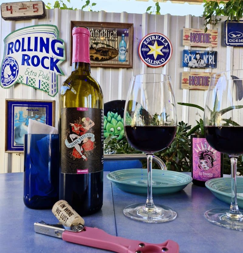 A bottle of La Dama Tatuada 2020 and glasses of red wine on a table at El Corcho Rosa, with La Traviesa behind